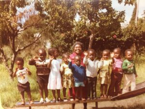 A smiling woman stands in front of a group of waving toddlers