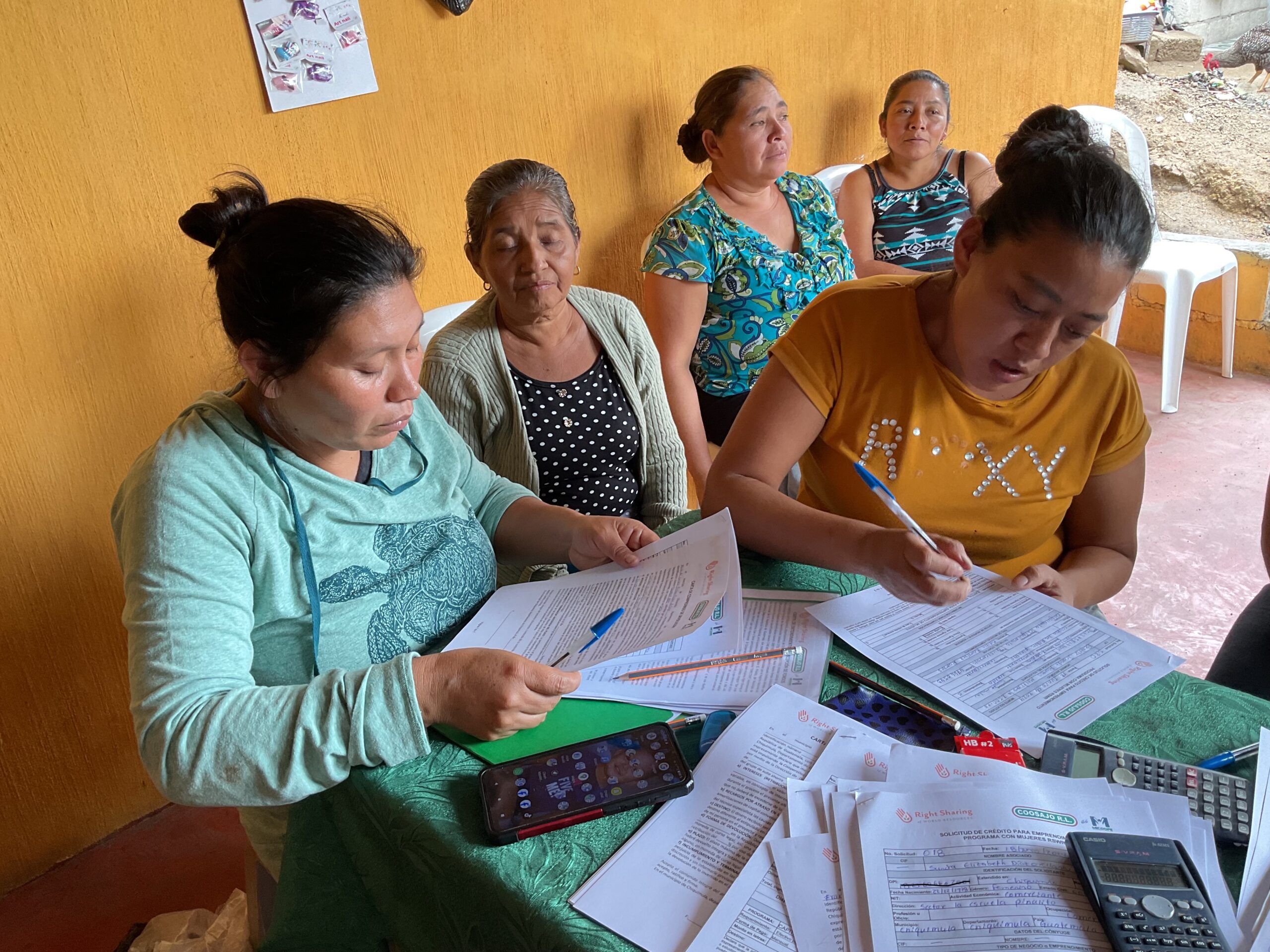 One woman completes paperwork while another woman reviews other papers. Three women sit behind them.