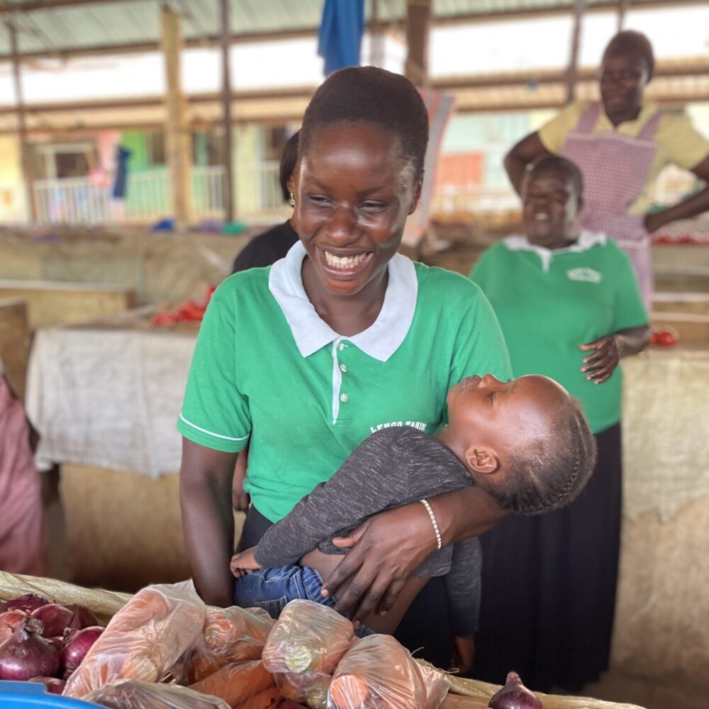 Lavinda with daughter in her arms, in front of vegetable stall