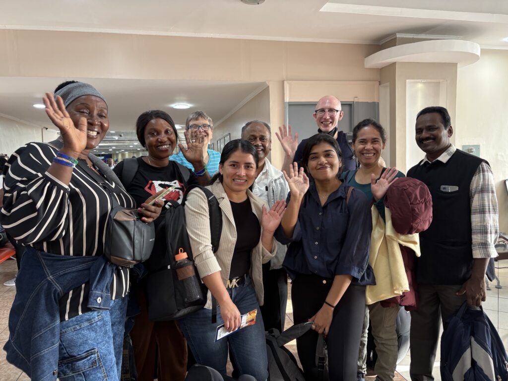 Nine people stand in an airport terminal waving at the camera. 