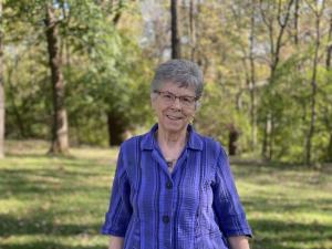A woman wearing a purple shirt stands in front of greenery