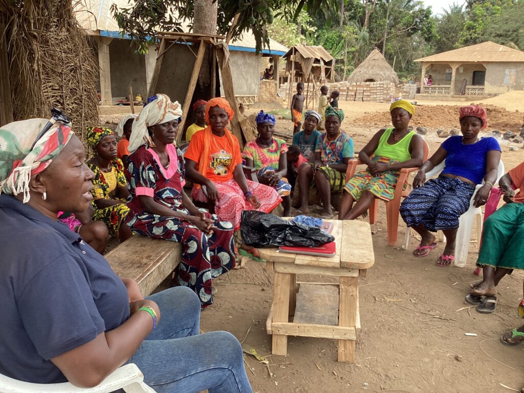 A woman wearing blue sits at the front of a group of ten women sitting in a semicircle 