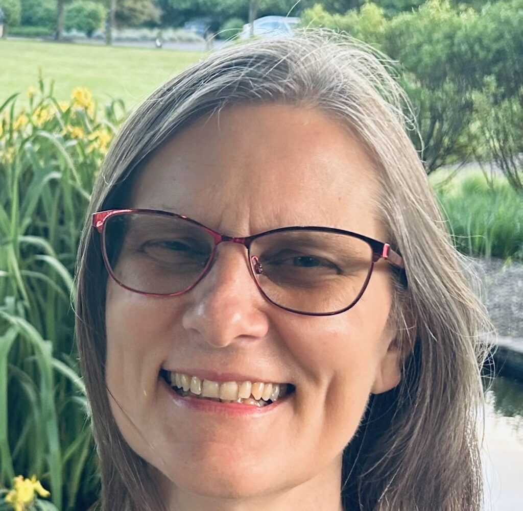 A woman wearing glasses stands in front of greenery