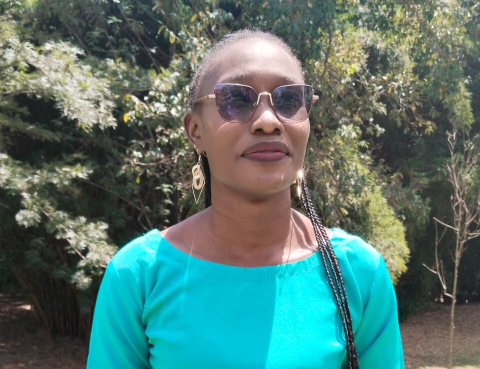 A woman wears a blue top and stands in front of greenery