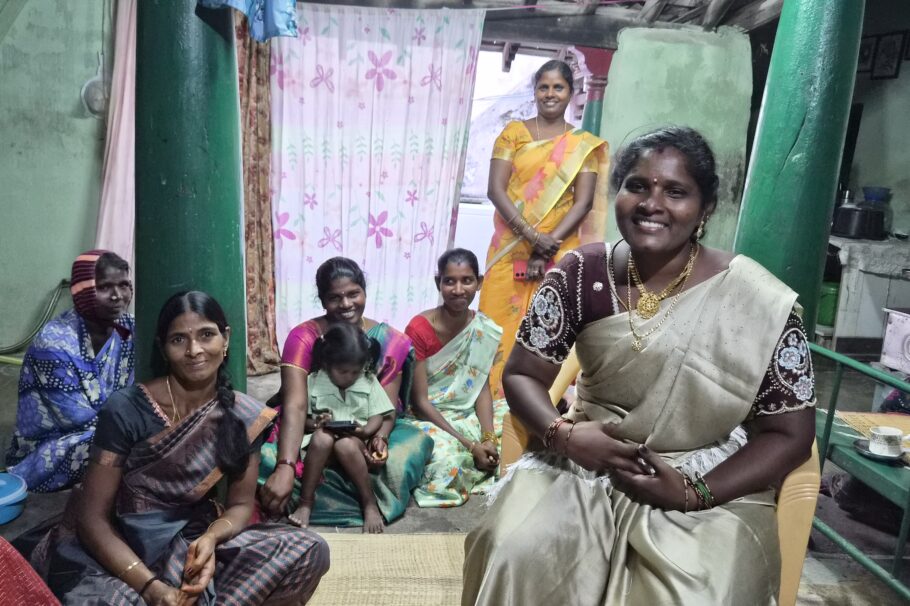 A woman wearing a beige sari sits on a chair in front of a group of women. The women are sitting on the floor behind her in a room with teal walls.