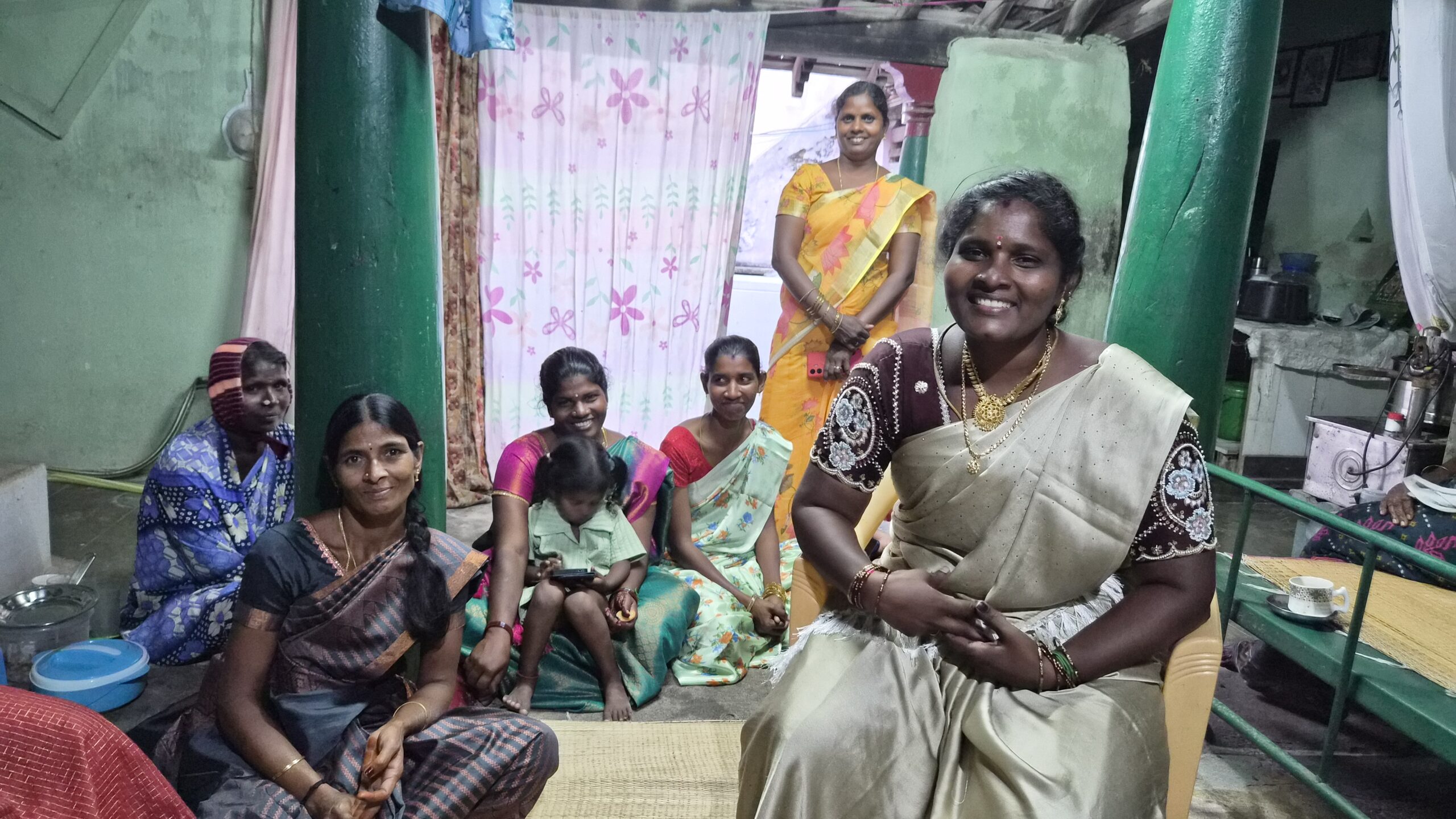 A woman wearing a beige sari sits on a chair in front of a group of women. The women are sitting on the floor behind her in a room with teal walls.