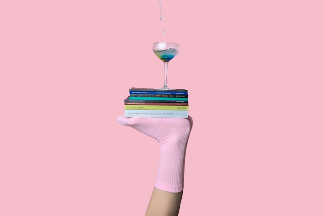 A glass rests atop of a stack of books being balanced on feet. The feet are covered in pink socks in front of a pink background.