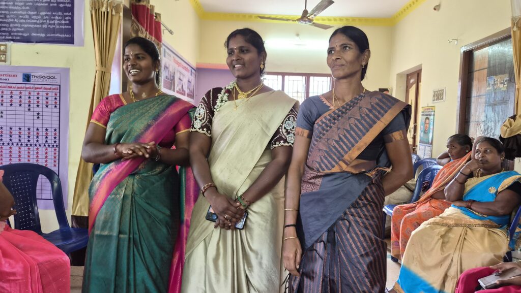Three women in saris stand in the middle of a yellow room. Other women sit in chairs along the wall.