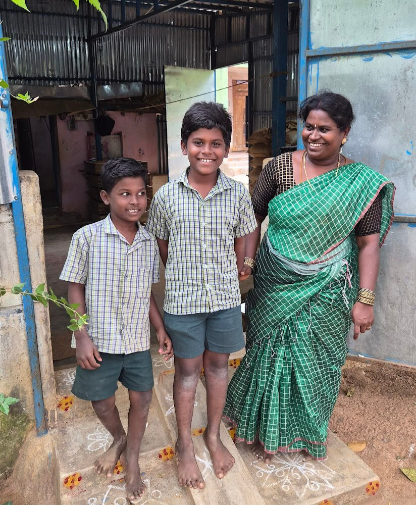 Two boys stand in front of a shop made of corrugated metal with their mother.