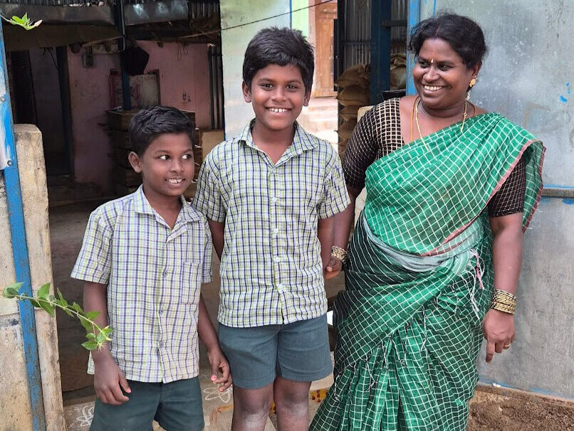 Two boys stand in front of a shop made of corrugated metal with their mother.