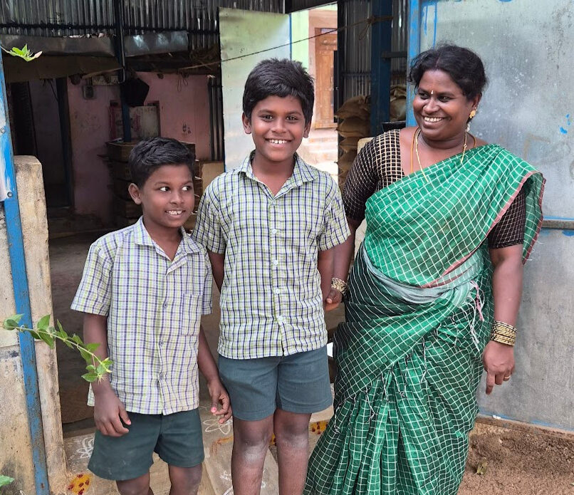 Two boys stand in front of a shop made of corrugated metal with their mother.