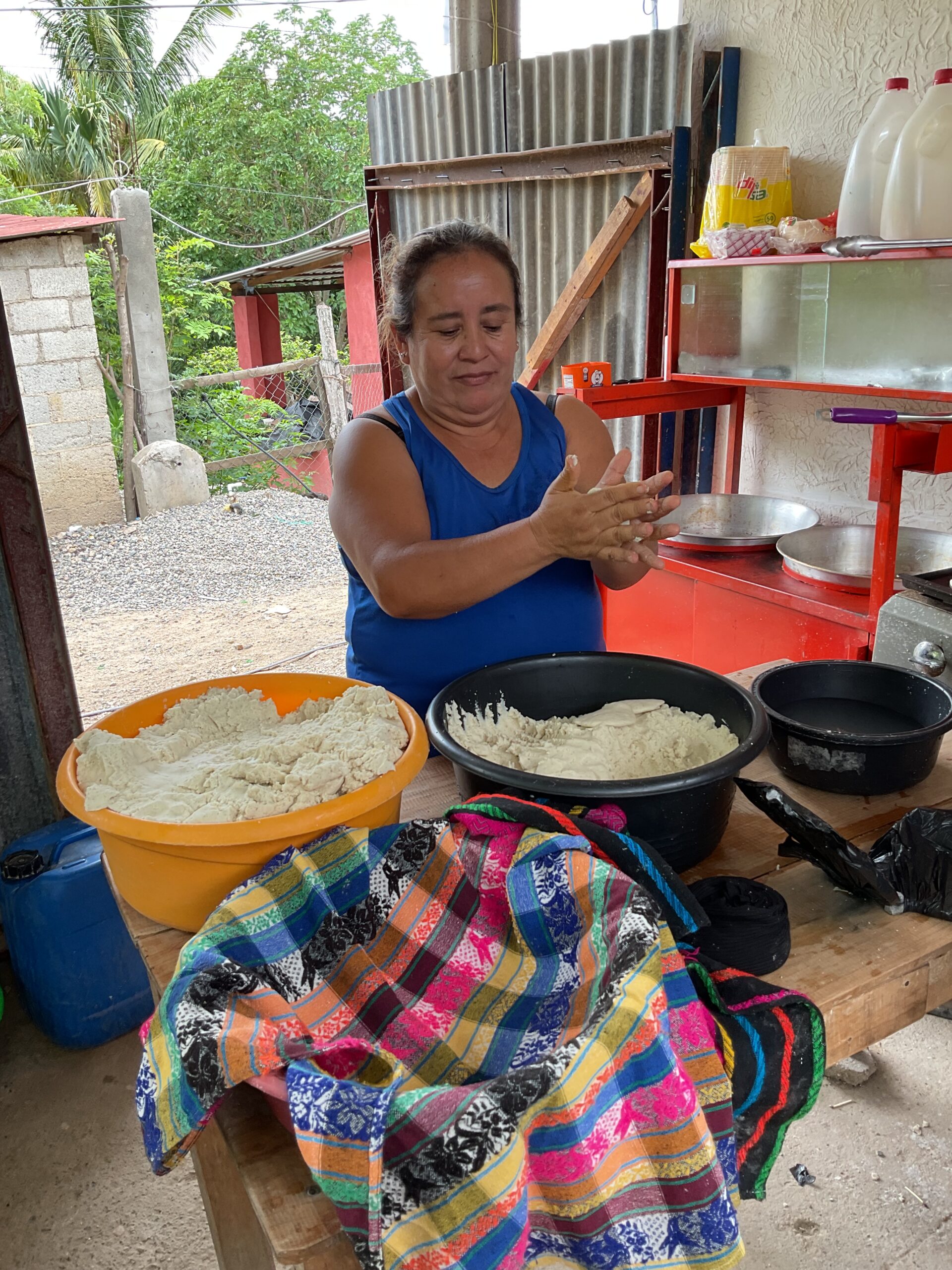 A woman stands in front of two large bowls of flour. She has a partially flattened tortilla in her hands.