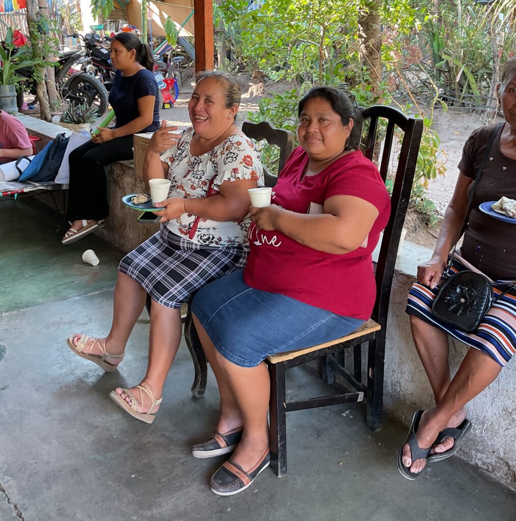 Two women sit together. They are sitting on chairs outside with cups of tea and plates of food in their hands.